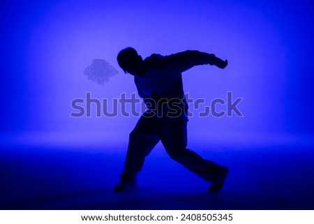 Silhouette of guy in casual clothes dancing elements of hip hop in studio with blue light. Dancer demonstrates body plasticity. Full height. Modern street choreography.