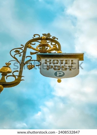 European style hotel sign with ornate structure. Sign reads, "Hotel Post". The sign post is adorned with grapevine elements. Set against a cloudy blue sky. Located in in old town Rüdesheim, Germany.