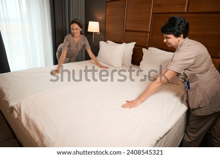 Two hotel maids busy with making bed in guest suite Royalty-Free Stock Photo #2408545321