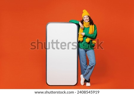 Full body young happy woman she wear green knitted sweater yellow hat scarf big huge point index finger on blank screen mobile cell phone smartphone with area isolated on plain orange red background