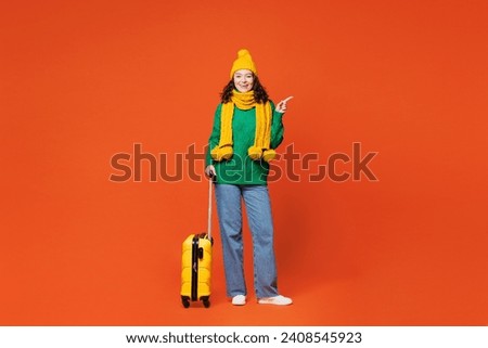 Traveler happy woman wear winter casual clothes hold bag point aside on area isolated on plain orange background. Tourist travel abroad in free spare time rest getaway. Air flight trip journey concept
