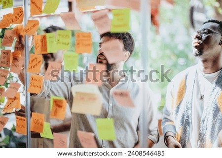 Diverse business partners collaborate in a modern office, brainstorming and planning with post-it notes on a glass wall. Successful teamwork and cooperation in achieving goals. Royalty-Free Stock Photo #2408546485