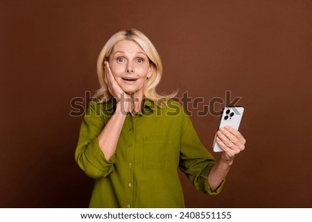 Photo of impressed woman with blond hair dressed khaki shirt hold smartphone palm on cheekbone isolated on dark brown color background