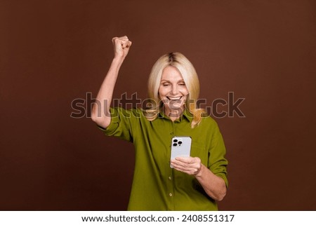 Photo of overjoyed woman with blond hair dressed khaki shirt look at smartphone win lottery isolated on dark brown color background