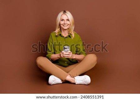 Full size photo of smart positive senior woman dressed khaki shirt sit on floor holding smartphone isolated on dark brown color background