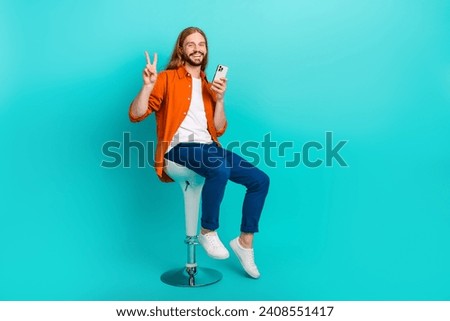 Full size photo of cheerful guy dressed stylish outfit sit on stool hold smartphone show v-sign isolated on turquoise color background