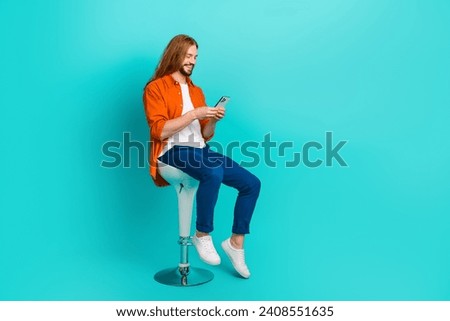 Full size photo of handsome guy dressed blue jeans sit on stool read notification on smartphone isolated on turquoise color background