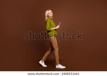 Full size photo of nice woman with blond hair dressed khaki shirt hold smartphone go empty space isolated on dark brown color background