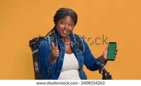 African american bike messenger vertically holding mobile device with isolated green screen display. Female courier showing delivery app on smartphone with chromakey mockup template.
