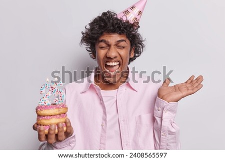 Anniversary celebration concept. Overjoyed Hindu man with curly hair celebrates 25th birthday exclaims loudly keeps palm raised up wears cone party hat and pink shirt poses against white background.