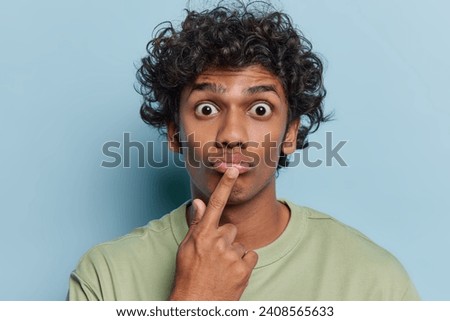 Portrait of Hindu man with curly hair keeps finger on lips stares bugged eyes with big surprisement reacts to something shocking isolated over blue background. Human facial expressions and reactions