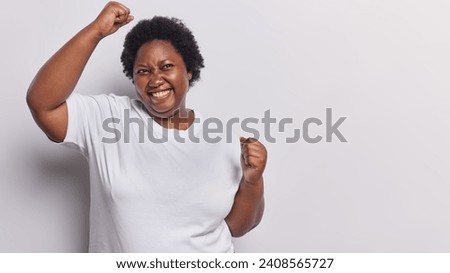 Photo of overjoyed dark skinned plump woman makes winner gesture raises arms and clenches fists feels like winner dressed in casual t shirt isolated over white background copy space for your advert Royalty-Free Stock Photo #2408565727