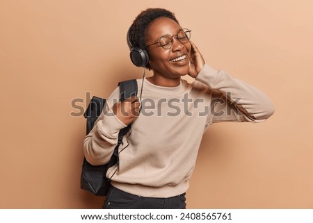 Happy joyful African girl returns home from school enjoys listening muic via headphones catches every bit has eyes closed dressed in casual sweatshirt carries rucksack isolated over brown background