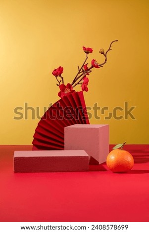 A pink podium, a paper fan, a peach blossom branch and a tangerine are artistically arranged on a red and yellow background. Display products with a festive atmosphere. Royalty-Free Stock Photo #2408578699