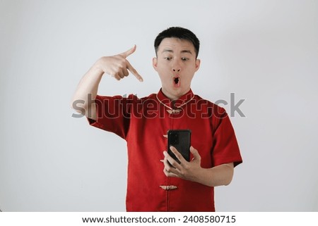 Surprised face of Asian man wearing Chinese traditional cloth or Cheongsam pointing his smartphone on white background Royalty-Free Stock Photo #2408580715