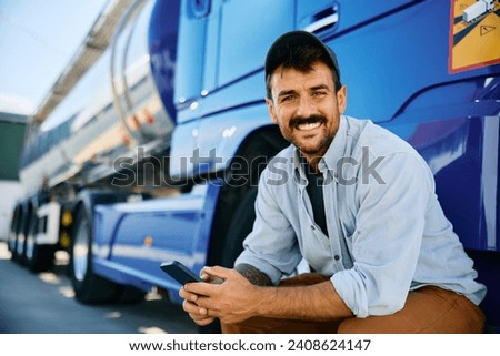Happy truck driver texting on mobile phone while taking a break on parking lot and looking at camera. Copy space.  Royalty-Free Stock Photo #2408624147
