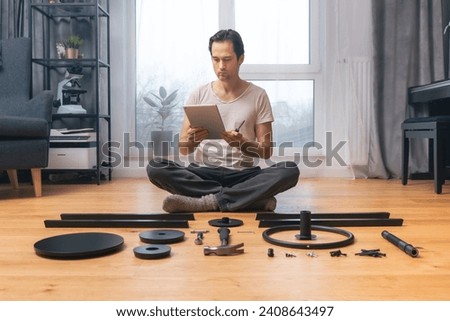 A man on a tablet is watching a video tutorial on self-assembly of furniture at home, sitting in a room on the floor among a set of neatly laid out parts and tools. Video instructions for beginners. Royalty-Free Stock Photo #2408643497