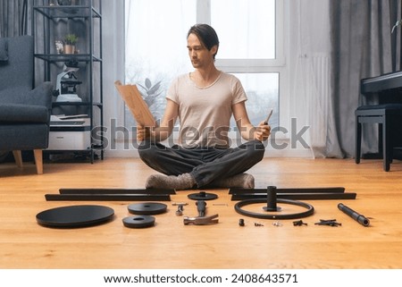 A perfectionist man carefully reads the instruction for assembling furniture, sitting on the floor in the lotus position, among neatly arranged parts and tools. Self-assembly of furniture from scratch Royalty-Free Stock Photo #2408643571