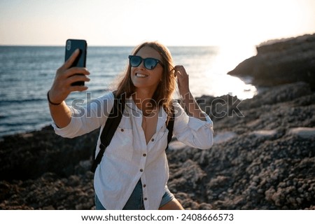 Happy traveler woman in white shirt and sungalsses taking selfie with mobile phone on the rocky shore at beautiful sunset