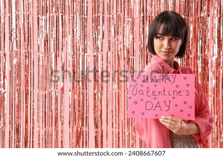 Beautiful young woman holding gift card with text HAPPY VALENTINE'S day and tinsel curtain on pink background Royalty-Free Stock Photo #2408667607