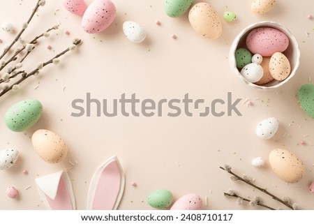 Celebrate Easter with this tender top view display. Bowl with color-packed eggs, whimsical bunny ears, pussy-willow twigs, sprinkles form cheerful ensemble on soft beige—ideal for your custom message Royalty-Free Stock Photo #2408710117