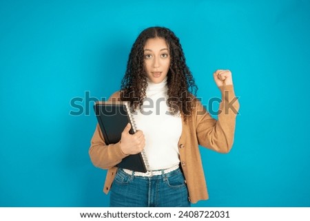 Shocked ecstatic Beautiful student girl holding books wearing formal clothes win luck lottery raise hands up shout yea