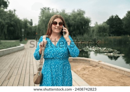Young pretty woman model in stylish dresses talk by mobile cell phone look aside standing on summer in spring park outdoors. Urban lifestyle concept. High quality photo Royalty-Free Stock Photo #2408742823