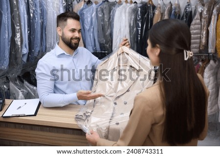 Dry-cleaning service. Happy worker giving coat to client indoors Royalty-Free Stock Photo #2408743311