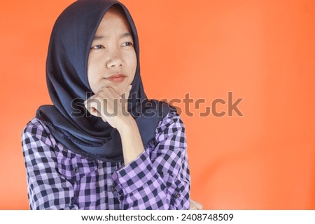 Close Up photo of stunning asian woman in black hijab looking in empty space deep thinking creative person arms on chin wearing purple flannel shirt isolated orange color background