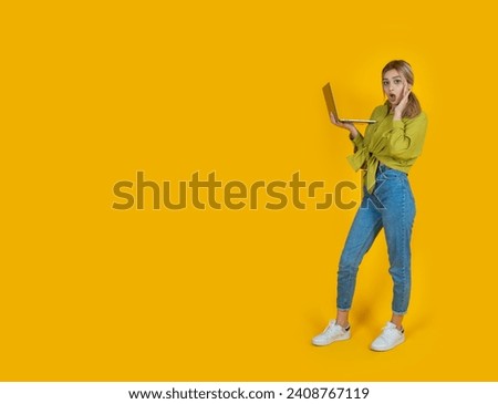 Shocked young woman, full body portrait of shocked young woman. Blonde girl hold use laptop amazed surprised isolated over studio yellow background copy space. Open mouth touching her face.