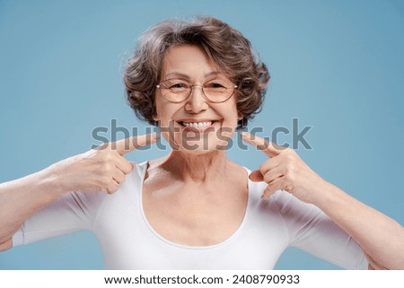 Portrait of smiling beautiful mature woman wearing eyeglasses pointing fingers at teeth, isolated on blue background. Concept of dental care, treatment Royalty-Free Stock Photo #2408790933