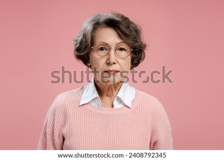 Portrait of serious, gray haired senior woman, grandmother, wearing eyeglasses, looking at camera standing isolated on pink background. Advertisement concept