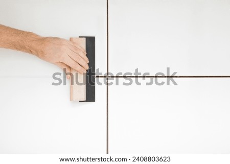 Young adult man hand using rubber trowel and grouting seams with paste between white ceramic tiles on wall or floor. Closeup. Renovation process. Repair work of home. Royalty-Free Stock Photo #2408803623