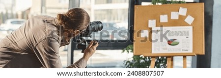 office manager in casual clothes adjusting digital camera near corkboard with infographics, banner