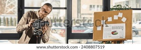 manager in casual wear and eyeglasses adjusting digital camera next to corkboard with graphs, banner