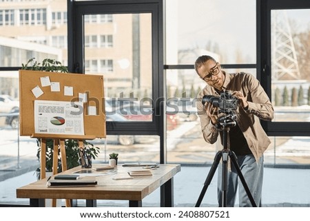 businessman in eyeglasses adjusting digital camera near desk and corkboard with charts in office