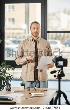 serious businessman holding document and talking in front of digital camera in modern office