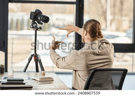 back view of businessman gesturing in front of digital camera at workplace in office, video blogger
