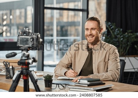 happy businessman smiling at digital camera and writing in notebook at workplace in modern office