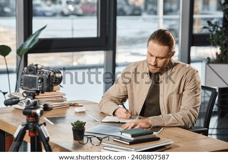 serious businessman in casual attire writing in notebook at work desk in front of digital camera