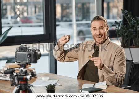 cheerful businessman showing bitcoins while sitting at workplace in front of digital camera