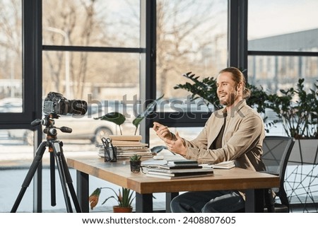 joyful businessman talking and gesturing in front of digital camera at work desk with notebooks