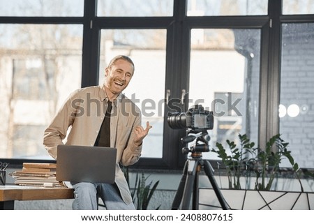 cheerful businessman with laptop talking and gesturing near digital camera in modern office