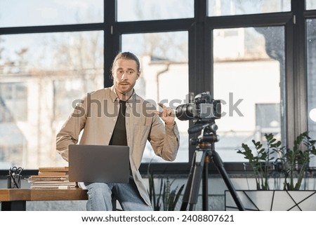 creative businessman with laptop talking and gesturing in front of digital camera in office