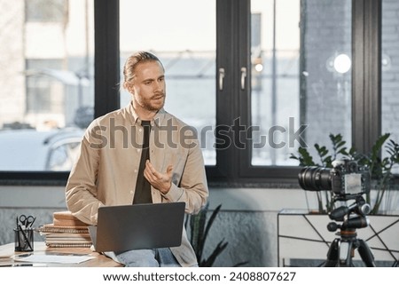 serious businessman with laptop talking in front of digital camera in office, video blogger