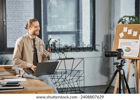 happy manager with laptop sitting on work desk in front of digital camera in contemporary office
