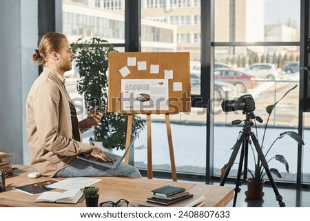 businessman with laptop sitting on work desk near digital camera and flip chart in modern office