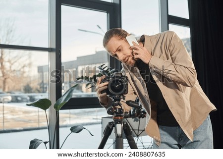 businessman talking on smartphone near professional digital camera in office, video blogger Royalty-Free Stock Photo #2408807635