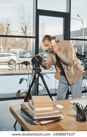 businessman talking on smartphone and adjusting digital camera next to work desk in modern office