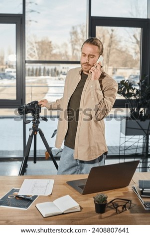 businessman talking on smartphone near digital camera and laptop on work desk in modern office